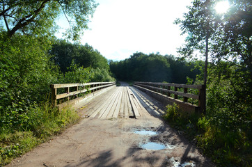 landscape, old wooden bridge over the river among the trees on a summer sunny day, puddles on a sandy road, clear blue sky with clouds and bright sun
