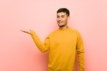 Young hispanic man showing a copy space on a palm and holding another hand on waist.