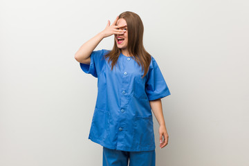 Young nurse woman against a white wall blink at the camera through fingers, embarrassed covering face.