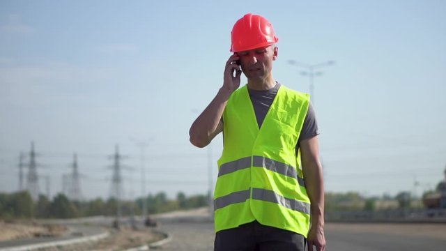 A worker in work clothes and a protective helmet walks and talks on the phone at a construction site at the construction site of the road on a summer sunny day. Slow motion