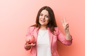 Young plus size curvy business woman holding a piggy bank showing victory sign and smiling broadly.