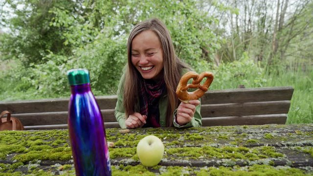 Girl sitting on wooden picknick beanch moss covered and looking through Brezel at the camera and smiling. Purple thermos and green apple in the foreground