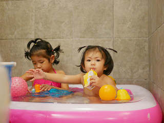 Little Asian baby girl (right) enjoys drinking milk and playing water, with her older sister, in a small pool in a bathroom at home - babies' leisure activity at home on a weekends