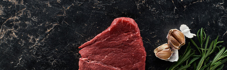 panoramic shot of garlic cloves, rosemary twigs and raw beef steak on black marble surface