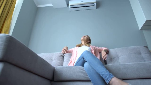 Young woman sitting on couch under air conditioner, cooling down on hot summer