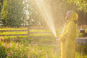 A young man in a yellow raincoat spraying a green lawn from a hose spraying water