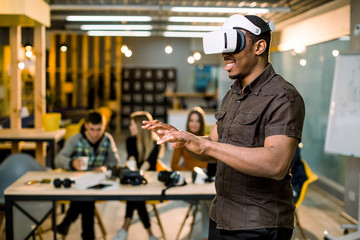 African young man wearing virtual reality glasses in modern interior design coworking studio. Smartphone using with VR goggles headset. Team of young people working at the table on the background - Powered by Adobe