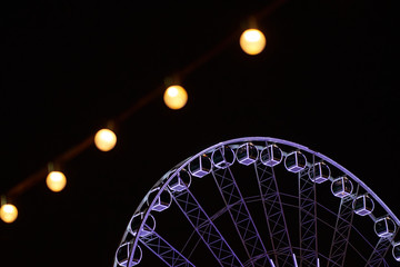 part of a ferris wheel at the fair, illuminated in white, in the foreground there are warm light bulbs out of focus