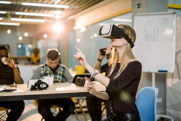 Young Caucasian woman in VR goggles at a desk with colleagues in an office