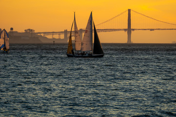 Two kissing sailboats below the Golden Gate Bridge at sunset.