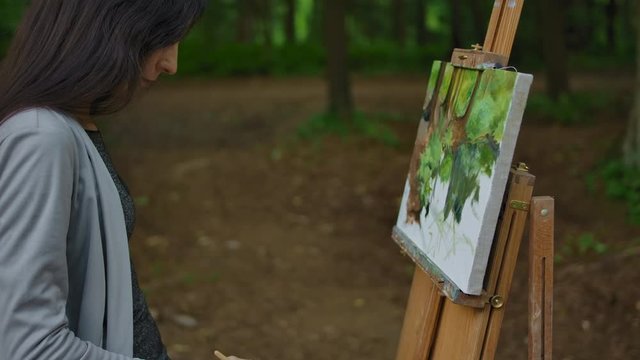 Female's hand painting a landscape on canvas in a park. Palm, brush, easel in the foreground, tree trunks of trees and greenery in the background. Stabilized side view in slow motion.