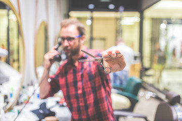 Modern man hairdresser holding sharp professional scissors and using telephone in barbershop.