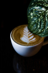 Cappuccino in white ceramic cup with latte art and large green leaf in cup from the flower. Fragrant drink in coffee shop on black background.