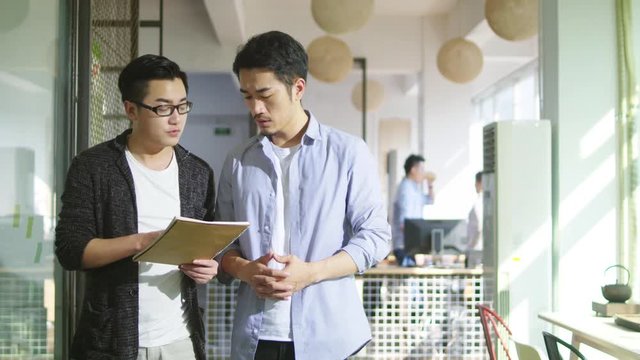 two young asian business men discussing sales report while walking in office