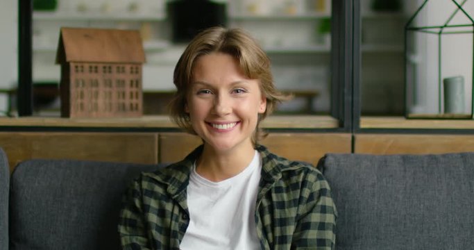 Portrait of young cheerful woman sitting on sofa, laughing at camera, looking happy and successful, in positive mood, short straight brown hair, grey eyes, Caucasian. 4K, shot on RED camera.