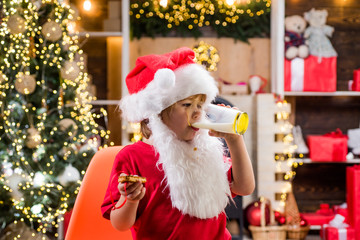 Fototapeta na wymiar Kid Santa Claus enjoying in served gingerbread cake and milk. Christmas for kid. Funny child Christmas.