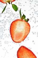 Sliced parts of a strawberry lie on a white background illuminated from above with water on it - background with fresh red strawberry with green on it in water with bubbles