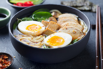 Asian noodle soup, ramen with chicken, tofu, vegetables and egg in black bowl. Slate background. Close up.