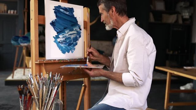 Bearded artist sitting in front of easel and painting abstract picture in studio