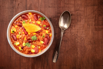 Chili con carne, a Mexican stew with red beans, cilantro leaves, ground beef, and chili peppers, top shot with a nacho chip on a dark rustic wooden background and a place for text