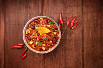 Chili con carne, a Mexican stew with red beans, cilantro, ground beef, and chili peppers, shot from the top with a nacho chip on a dark rustic wooden background