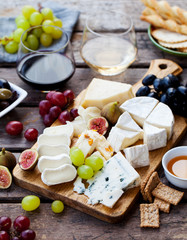 Cheese and fruits on cutting board with red, white wine on wooden background.