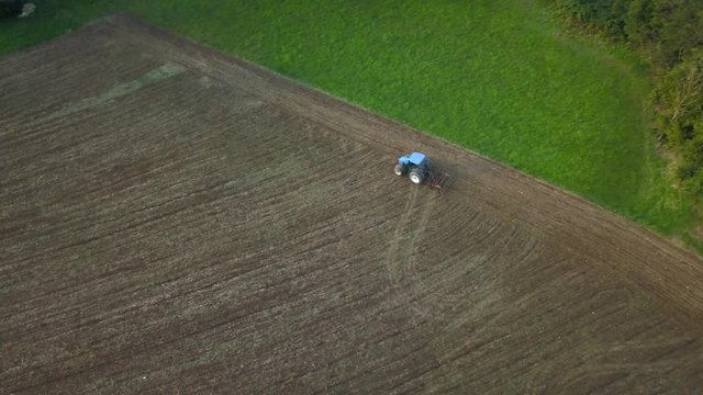 4K aerial view of blue farming tractor plowing the field next to the forest in the evening. stock video of the farmer ploughing the acre with a tractor