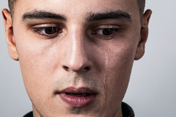 Obraz premium Unhappy crying young man with dropping tears on the face. Closeup studio portrait on grey background. Negative concept