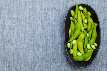 Fresh edamame green beans in black bowl. Grey textile background. Top view. Copy space.