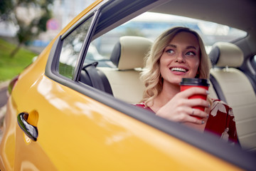 Image of happy blonde with glass of coffee in her hands sitting in back seat of yellow taxi.