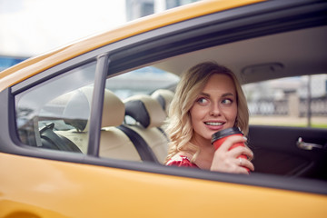 Image of curly blonde with glass of coffee in her hands sitting in back seat of yellow taxi