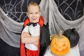 Portrait of funny cute little boy playing cheerfully in costume of vampire in beautiful spooky Halloween decor in black and white colors. Horizontal color photo.