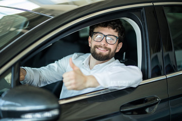 Happy handsome man smiling cheerfully showing thumbs up sitting in a new auto at the dealership salon copyspace car rental retail sales offer discount customer buyer consumerism.