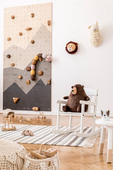 Stylish interior design of childroom with modern climbing wall for kids, design furnitures, soft toys, teddy bear on armchair and cute children's accessories. Scandinavian playroom decor. Template.