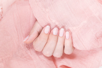 Closeup view of one beautiful female hand isolated on soft pink fabric texture background. Fingers with fresh pastel nude look manicure at short oval nails. Trend of natural beauty concept.