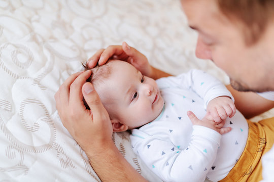 Close Up Of Handsome Young Dad Cuddling And Talking To His Adorable Six Months Old Son. Baby Looking At Father While Lying In Bed.