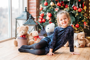 Obraz na płótnie Canvas Christmas fun. Happy little girl sitting on floor, smiling. Blur window, decorated fir tree and teddy bears in background.