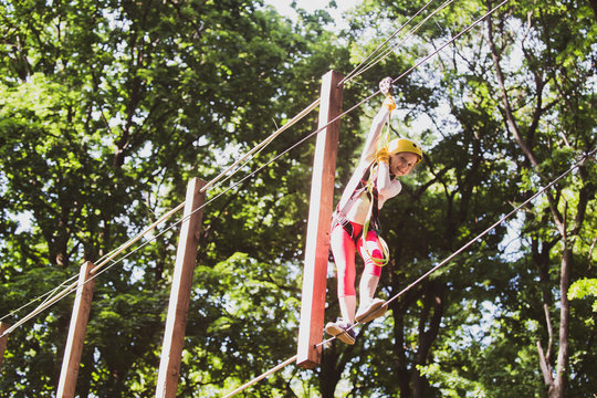 Cute little girl in climbing safety equipment in a tree house or in a rope park climbs the rope. Happy little girl calling while climbing high tree and ropes. Little girl concept.