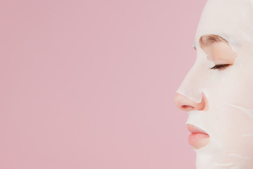 Beautiful young woman is applying a cosmetic tissue mask on a face on a pink background. Healthcare and beauty treatment and technology concept