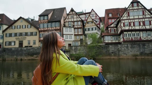 Beautiful woman in yellow coat with backpack sits near water. Girl closed her eyes in pleasure. Traditional half-timbered houses of Germany with canal reflections are on the background. Relax time