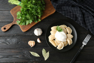 Delicious cooked dumplings with sour cream on dark wooden table, flat lay