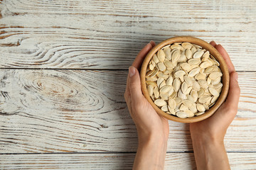 Young woman with bowl of raw pumpkin seeds at white wooden table, top view. Space for text