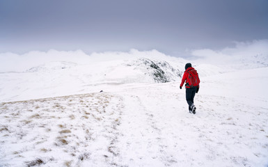 Fototapeta na wymiar A hiker in from the right edge of frame descending through snow from the summit of High Raise towards Rampsgill Head near Hartsop in the Lake District.