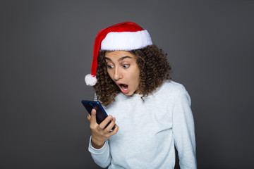 Girl in Santa Claus Christmas hat. A young brunette looks in surprise at the mobile phone. The concept of New Year's holiday promotions. Sell-out. 