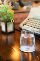 A glass of water with ice place on a dark brown wooden table with a paper coaster under a glass. there are an artificial flower and typewriter in the background