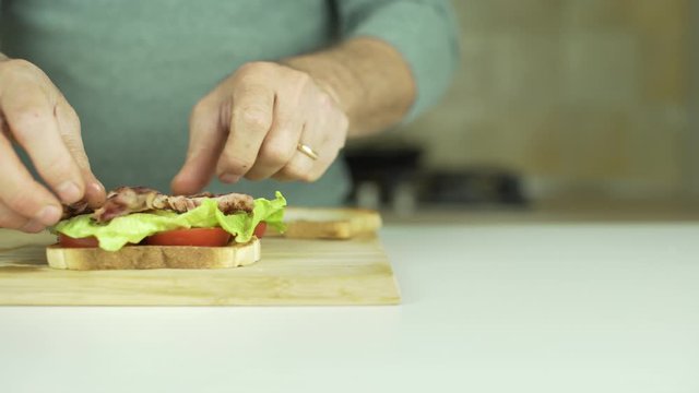 Dolly shot of man preparing a tasty BLT sandwich with red tomatoes, lettuce, bacon, then closing with sandwich loaf and press it with hands.