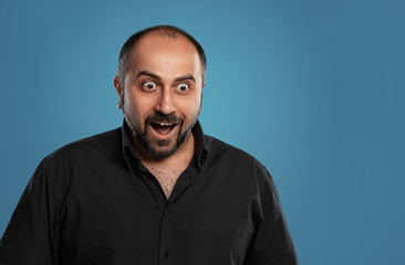 Obraz premium Close-up portrait of a brunet middle-aged man with beard, dressed in a black t-shirt and posing against a blue background.