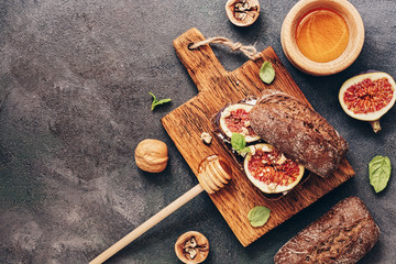 Fresh bread with soft cheese, figs, nuts and honey on a cutting board, dark rustic background. Top view, flat lay, copy space.