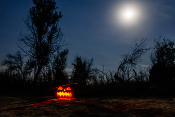Carved halloween pumpkin jack-o-lantern wearing witch hat with burning candles glows in a darkness. Spooky landscape