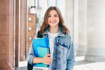 Young girl student smiling against university. Cute girl student holds folders and notebooks in...